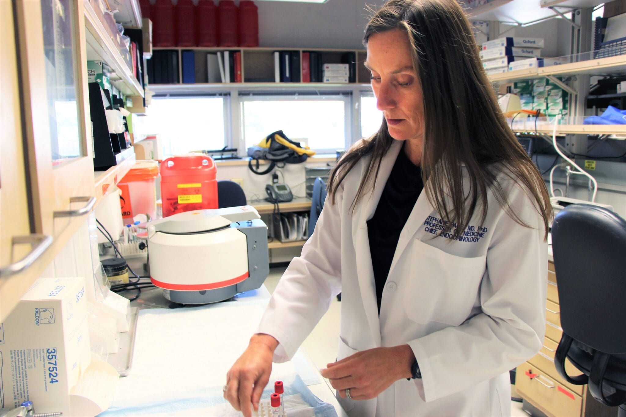 A woman in a white lab coat works at a laboratory bench, handling a small vial. Scientific equipment and supplies are visible on the shelves and counter around her.