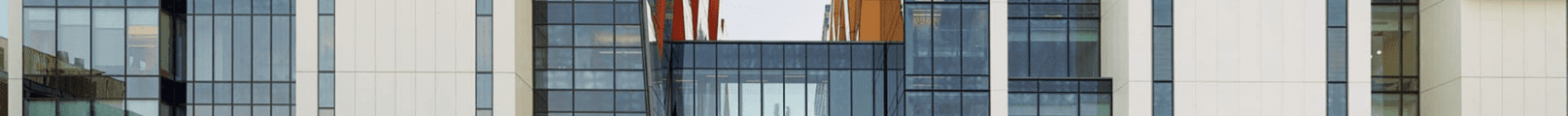 Modern building facade with vertical white panels and large glass windows, reflecting surrounding structures. Orange and red structural elements are visible behind the windows in the central section.