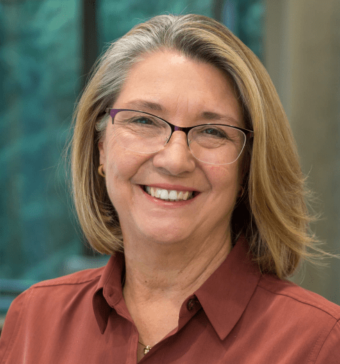 A smiling older woman with shoulder-length blonde hair and glasses, wearing a rust-colored shirt, stands indoors with a blurred background of large windows and greenery.
