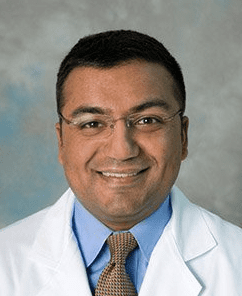 A man wearing glasses, a white medical coat, a blue shirt, and a yellow patterned tie smiles at the camera against a neutral, cloudy background.