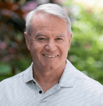 A smiling older man with short white hair wearing a light-colored, collared shirt stands outdoors in front of a blurred, green and leafy background.