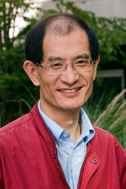 A middle-aged man with glasses, short black hair, and a receding hairline smiles while wearing a red jacket over a light blue shirt. Green foliage and a building are visible in the background.