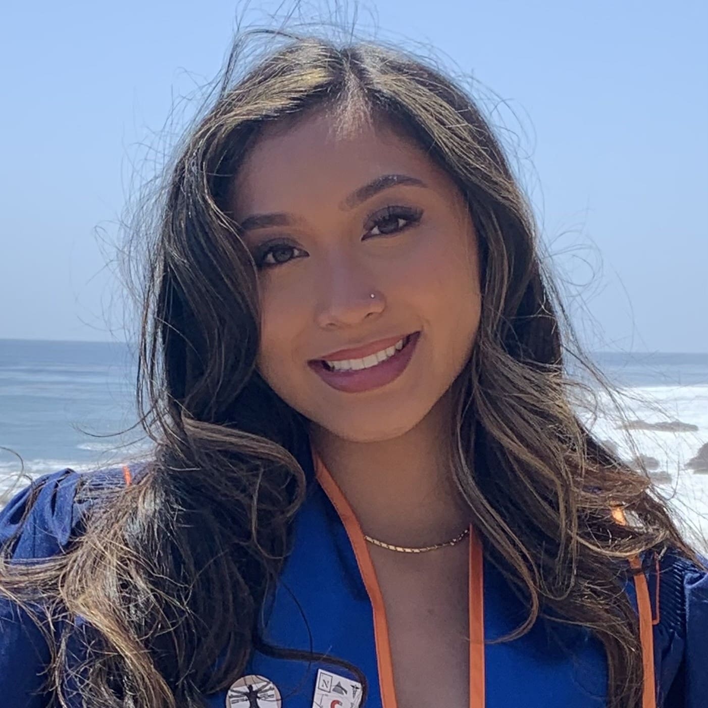 A young woman with long, wavy hair smiles at the camera, wearing a blue graduation gown with orange trim, standing outdoors by the ocean on a sunny day.