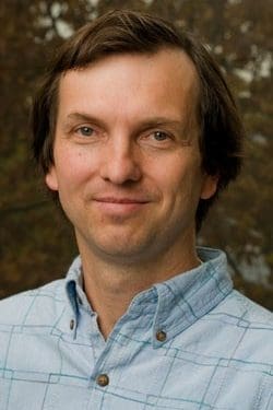 A man with light skin and straight brown hair is wearing a light blue, plaid button-up shirt. He is smiling softly and standing in front of a blurred, brownish outdoor background.