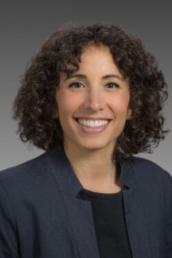 A woman with curly brown hair wearing a dark blazer and black top smiles at the camera against a gray background.