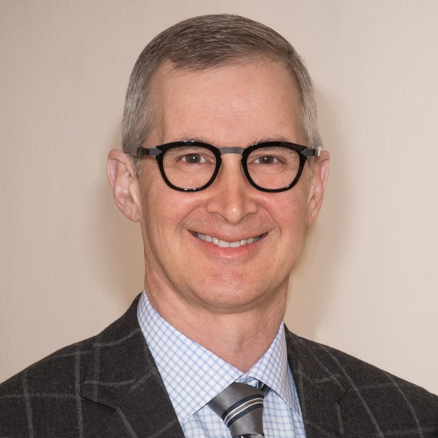 A man with short gray hair, wearing round black glasses, a checkered dress shirt, a striped tie, and a dark plaid suit jacket, smiling in front of a plain light-colored background.
