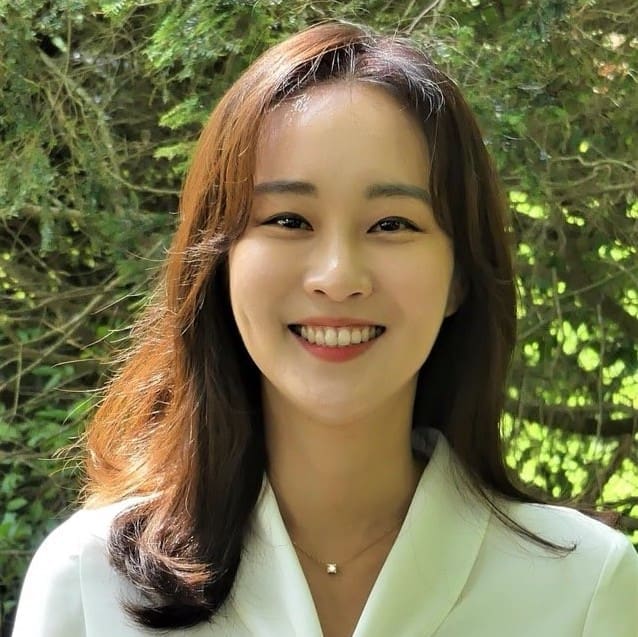 A young woman with long brown hair and a white top smiles warmly outdoors, with green foliage and sunlight in the background.