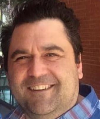 A man with dark hair and a beard smiles at the camera. He is wearing a blue and red plaid shirt, and there is a brick wall and some glass windows in the background.