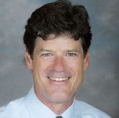 A man with short, curly dark hair wearing a light blue collared shirt and tie, smiling at the camera with a neutral, blurred gray background.