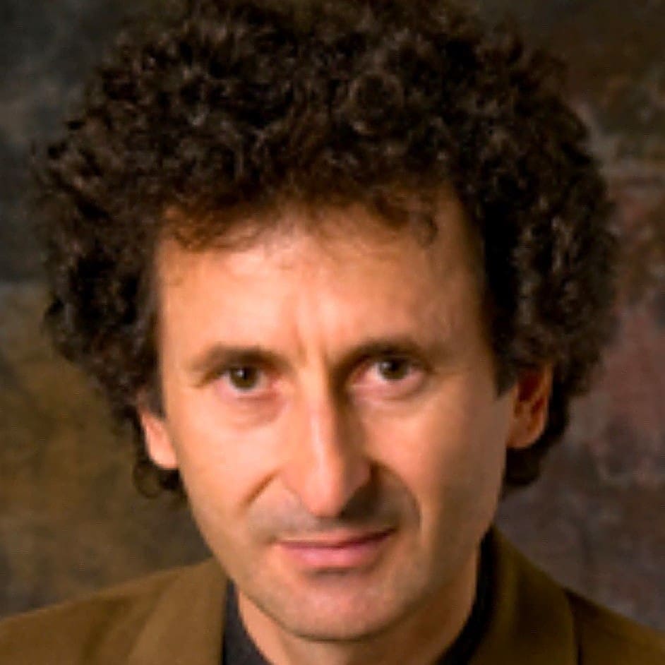 A man with curly dark hair wearing a brown jacket and a dark shirt, looking directly at the camera with a neutral expression. The background is softly blurred in brown and dark tones.