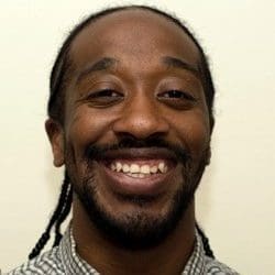 A man with braided hair and a beard smiles at the camera. He is wearing a patterned shirt and is positioned against a plain, light-colored background.