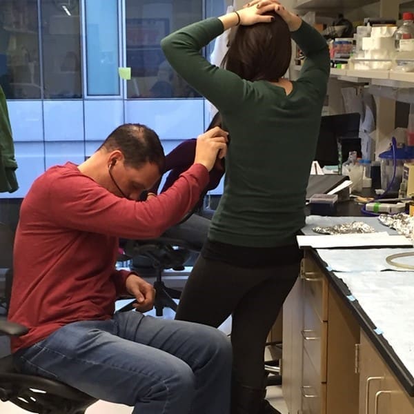 A man sitting on a lab chair examines a womans back as she stands with her hands on her head in a laboratory setting, with lab equipment and supplies visible on counters.