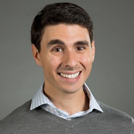 Smiling young man with short dark hair wearing a gray sweater over a light blue collared shirt, posing in front of a plain gray background.