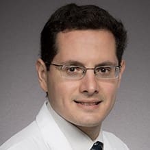 A man with short dark hair and glasses, wearing a white lab coat and a dark tie, smiles slightly in front of a plain gray background.