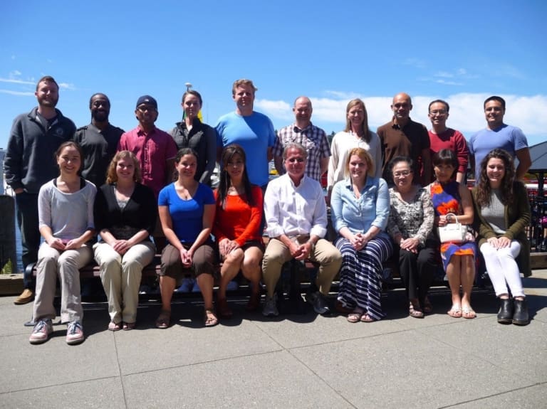 A group of seventeen people pose for a photo outdoors on a sunny day, with blue sky and water in the background. Eight people are seated in front, while nine stand behind them, all smiling at the camera.