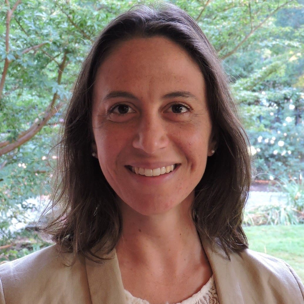 A woman with long brown hair smiles at the camera outdoors, with green trees and plants in the background. She is wearing a light-colored blazer and a white top.