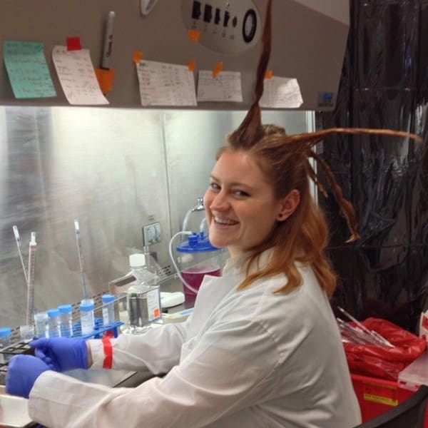 A woman in a lab coat and gloves smiles while working at a lab bench. Her hair is styled into several spiky points sticking out in different directions. Lab equipment and notes are visible around her.