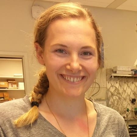 A smiling person with long blonde hair in a braid, wearing a gray shirt, standing in a well-lit room with shelves and equipment in the background.