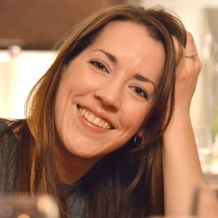 A woman with long brown hair smiles warmly at the camera, resting her head on her hand. She is wearing a gray top and a ring on her finger. The background is softly blurred.