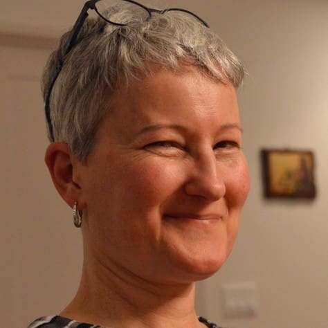 A person with short gray hair and glasses resting on their head is smiling with one eye closed. They are wearing hoop earrings and standing indoors with a blurry painting in the background.
