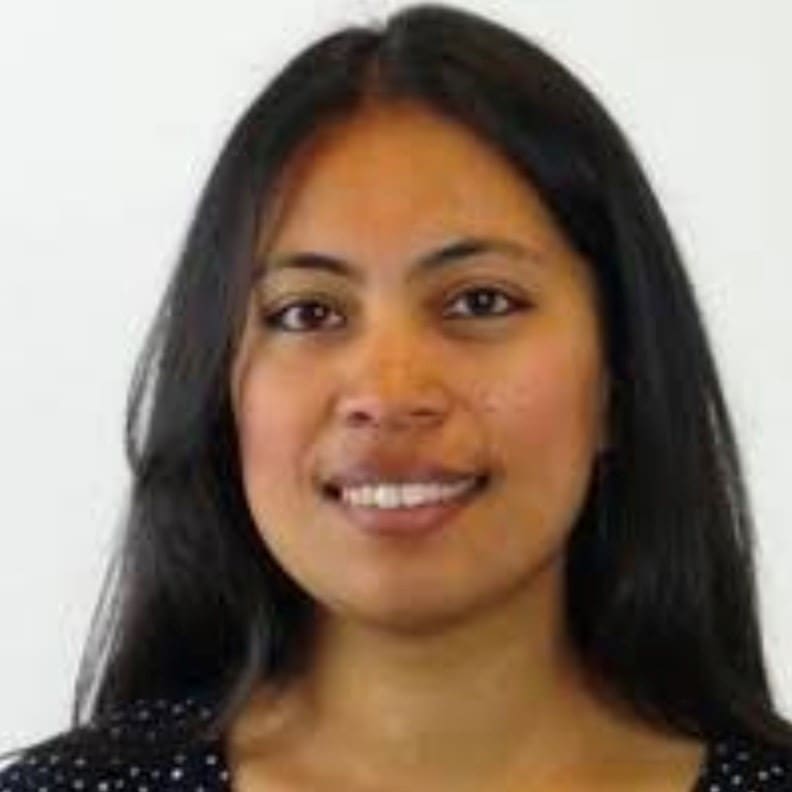 A woman with long straight dark hair smiles slightly at the camera. She is wearing a black top with small white dots and is pictured against a plain white background.