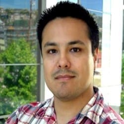 A man with short dark hair and a light beard wears a red and white plaid shirt, standing indoors near large windows with greenery and buildings visible outside.