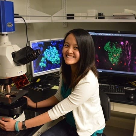 A woman sits at a microscope in a lab, smiling at the camera. Behind her, two monitors display colorful cellular images, likely from her research, and the desk is filled with lab equipment.