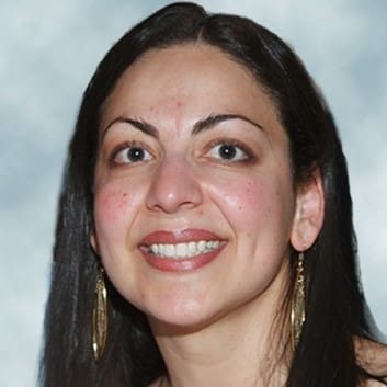 A woman with long dark hair, wearing gold dangling earrings, smiles at the camera against a soft, blurred background.