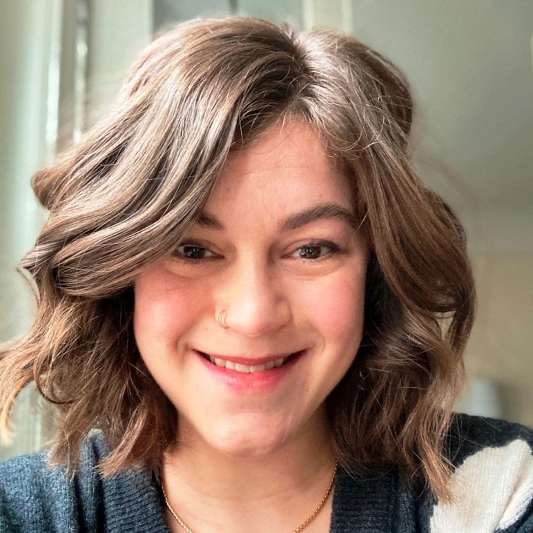A person with wavy brown hair and a nose ring smiles at the camera in a well-lit indoor setting. They are wearing a dark top and a gold necklace.