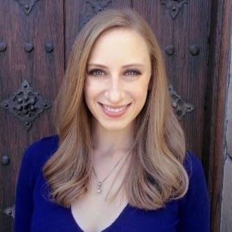 A woman with long, straight blonde hair and blue eyes is smiling at the camera, standing in front of a wooden door with ornate metal details. She is wearing a blue top and a necklace.