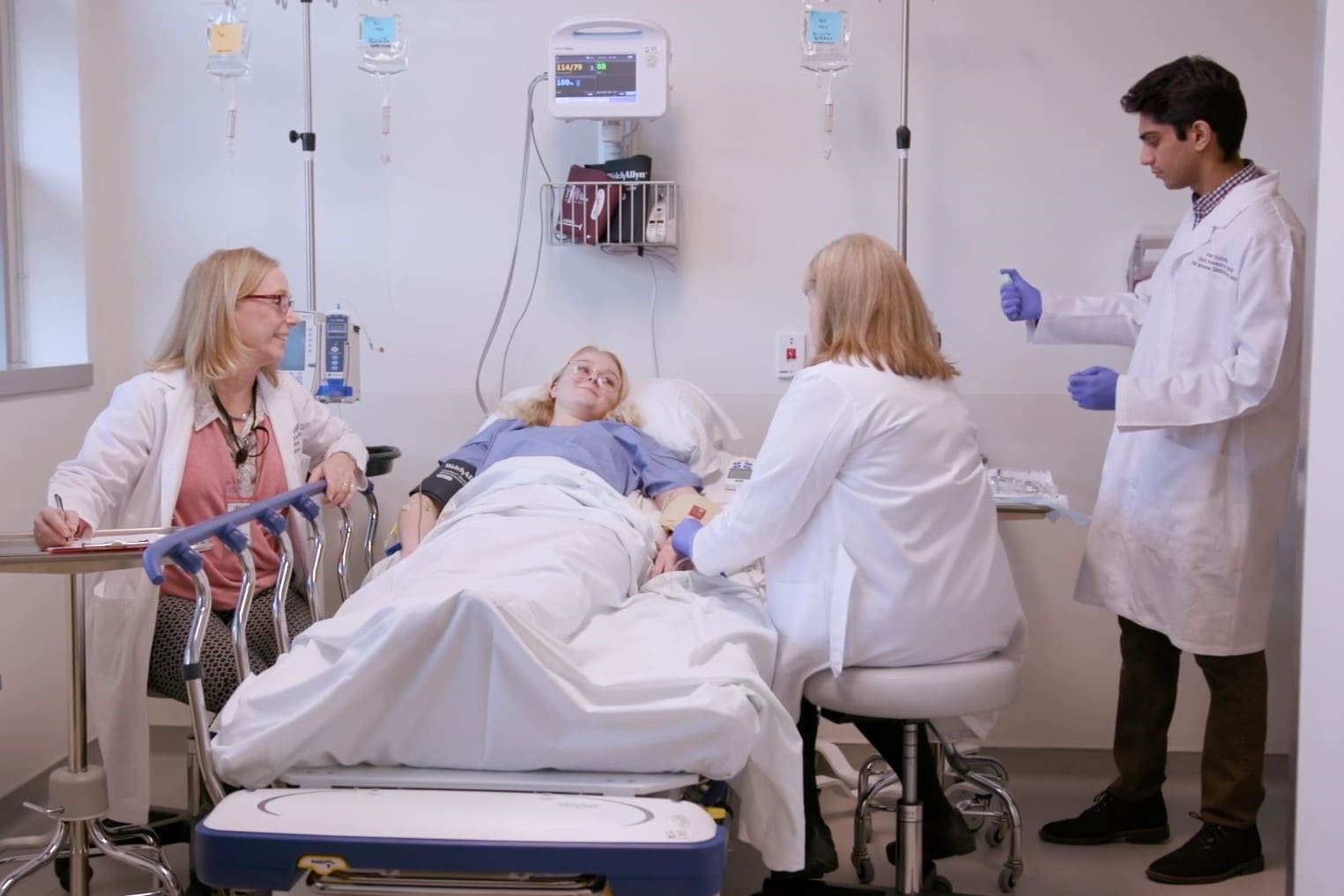 A patient lies in a hospital bed surrounded by three medical professionals. One stands holding a syringe, while the others sit nearby, monitoring and talking to the patient. Medical equipment is visible in the room.