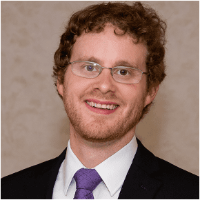 A man with curly brown hair, glasses, and a short beard is smiling. He is wearing a black suit, white shirt, and purple tie, standing in front of a plain, light-colored background.