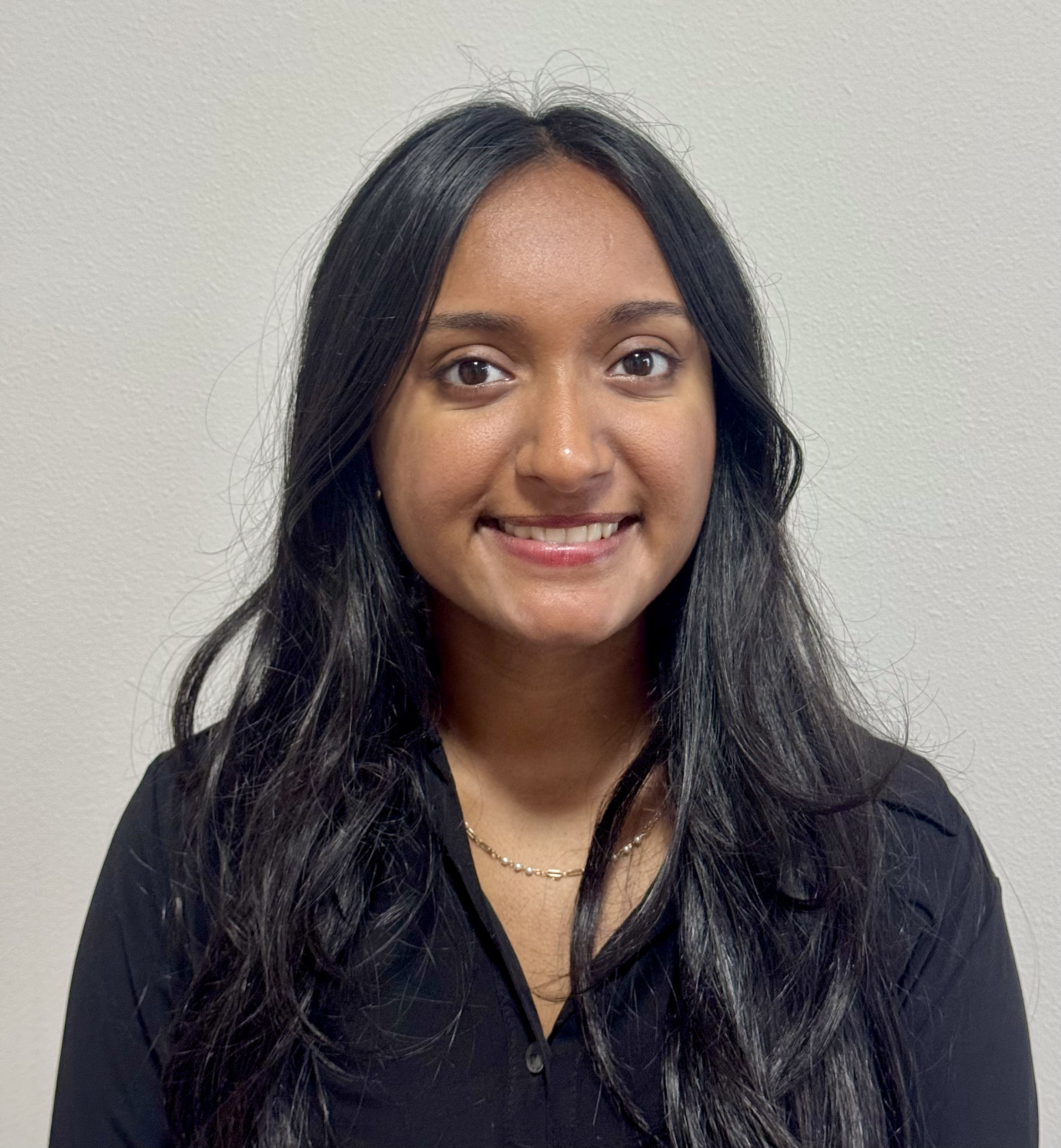A woman with long, straight black hair and medium skin tone smiles at the camera. She is wearing a black top and a delicate necklace, standing in front of a plain light-colored wall.