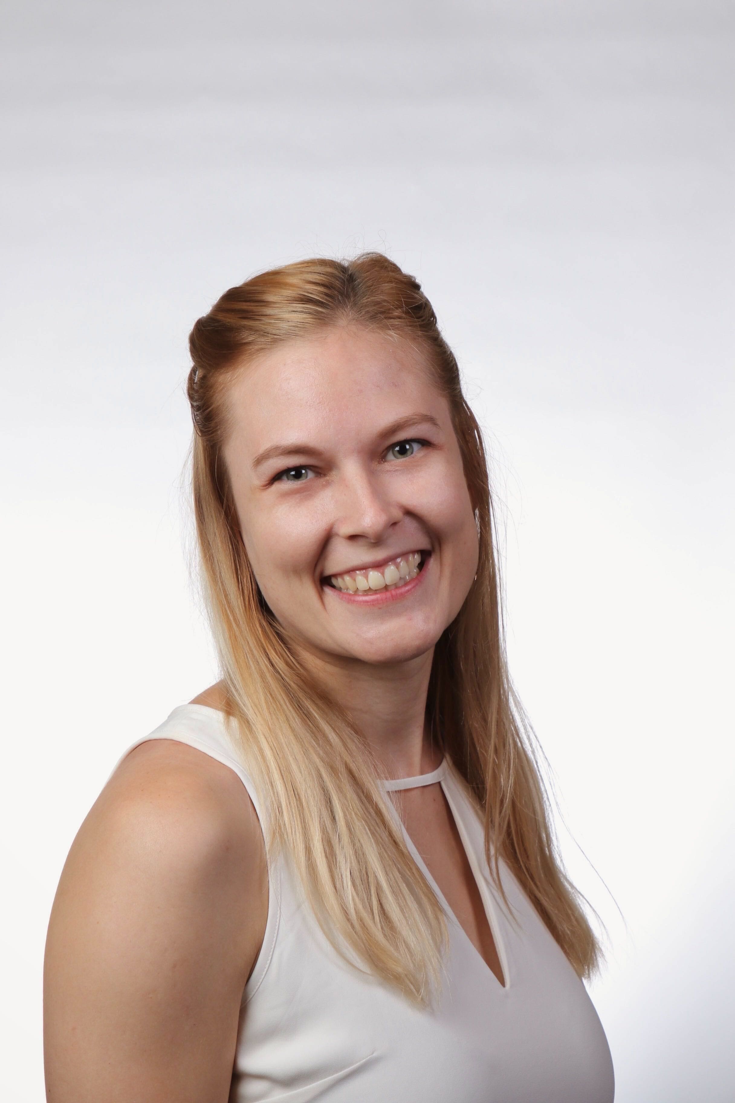 A young woman with long blond hair and a white sleeveless top smiles at the camera against a plain white background.