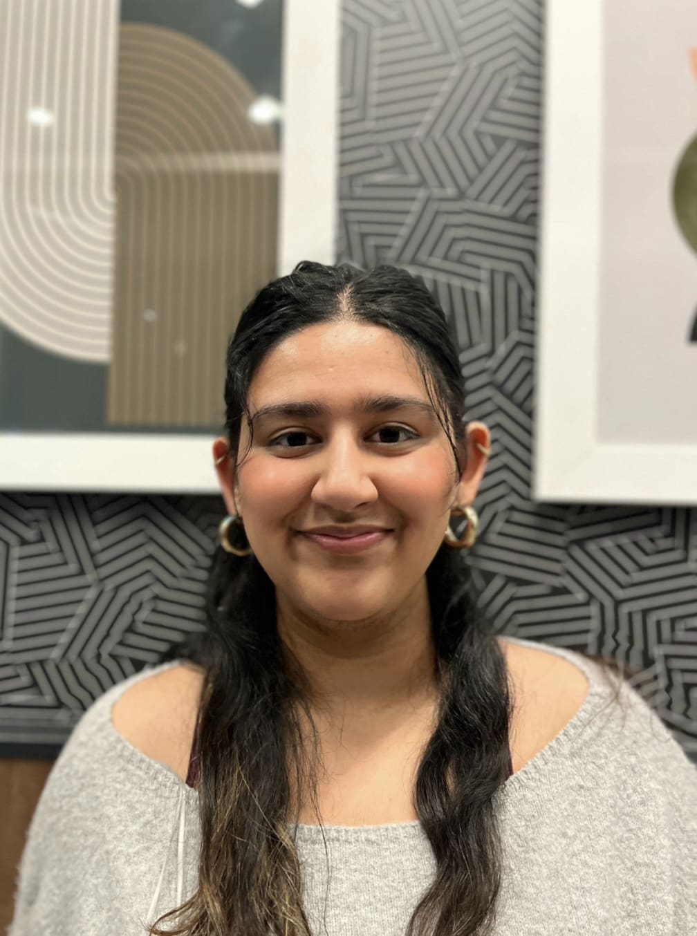 A young woman with long dark hair, wearing large hoop earrings and a light gray off-the-shoulder top, smiles in front of a modern, geometric patterned wall with framed abstract art.
