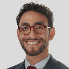 A man with curly dark hair, a trimmed beard, and mustache, wearing round red glasses, a dark suit, a light blue shirt, and a red tie, smiling at the camera against a light gray background.