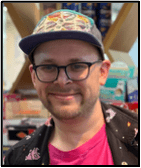 A smiling person wearing glasses, a colorful cap, and a black jacket with a pink shirt stands indoors in front of store shelves filled with various products.