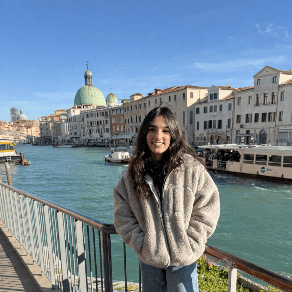 A young woman with long dark hair wearing a gray fleece jacket stands smiling by a canal in Venice, Italy, with historic buildings and a dome visible in the background under a clear blue sky.