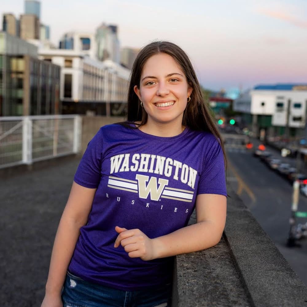 A young woman smiles while standing on an urban overpass at sunset, wearing a purple Washington Huskies t-shirt. City buildings and a street with traffic are visible in the background.