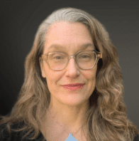 A woman with long, wavy gray hair and glasses smiles slightly. She is wearing a black top and a light blue pendant necklace, with a neutral dark background behind her.