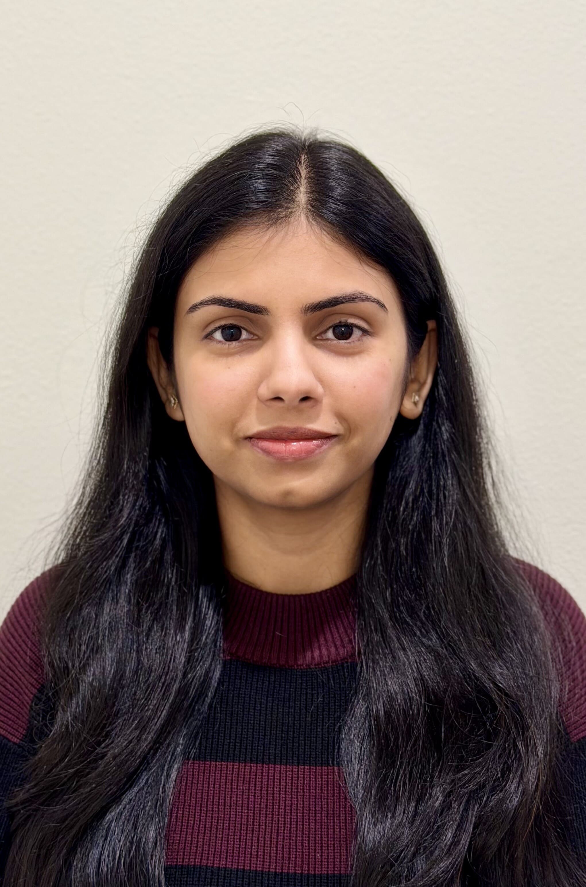 A woman with long, straight black hair is facing the camera, wearing a black and maroon striped sweater. She has a neutral expression and is standing in front of a plain light-colored wall.