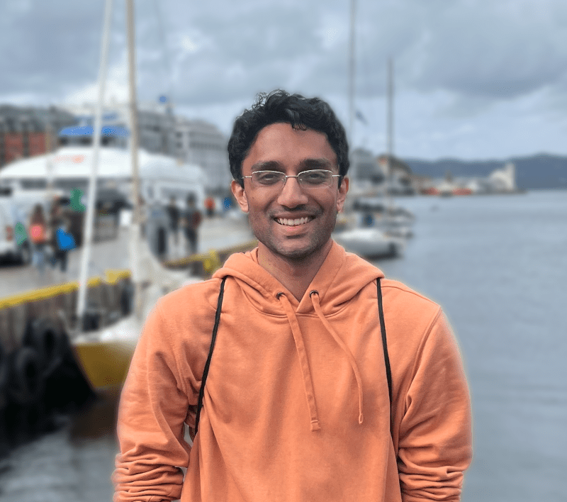 A smiling person wearing glasses and an orange hoodie stands by a waterfront, with boats, water, and cloudy skies in the background.